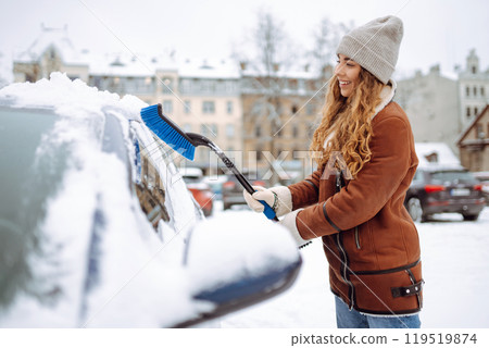 Portrait of young woman cleaning snow off car during winter snowfall. Winter, vehicle concept. Portrait of young woman cleaning snow off car during winter snowfall. Winter, vehicle concept. 119519874