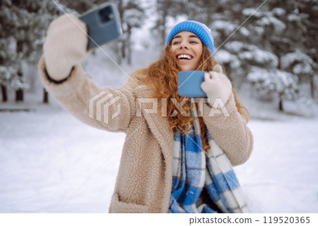 Portrait of a beautiful woman in warm clothes takes selfie with a hot drink from a thermos in hands. Portrait of a beautiful woman in warm clothes takes selfie with a hot drink from a thermos in hands. 119520365