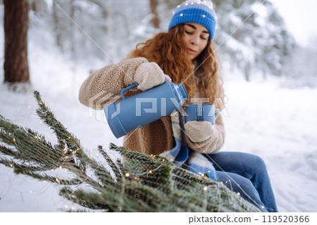 Young woman drinks a hot drink from a thermos while sitting in a snowy forest with a Christmas tree. Young woman drinks a hot drink from a thermos while sitting in a snowy forest with a Christmas tree. 119520366