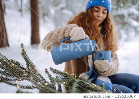 Young woman drinks a hot drink from a thermos while sitting in a snowy forest with a Christmas tree. Young woman drinks a hot drink from a thermos while sitting in a snowy forest with a Christmas tree. 119520367