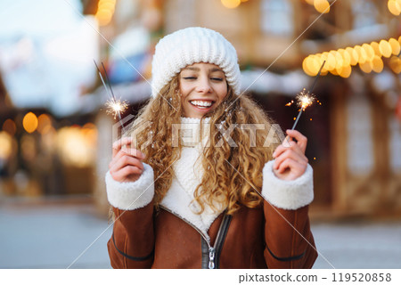 Cheerful young woman holding a sparkler in hand in the winter street. Festive garland lights. 119520858