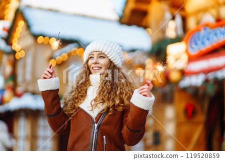 Cheerful young woman holding a sparkler in hand in the winter street. Festive garland lights. 119520859