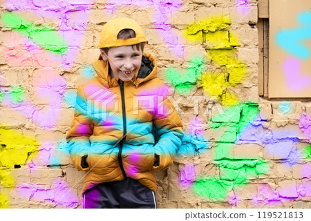 Child in graffiti. The boy is standing against a brick wall in fashionable clothes and a baseball cap, outlined with bright colors. Child in graffiti. The boy is standing against a brick wall in fashionable clothes and a baseball cap, outlined with bright colors. 119521813