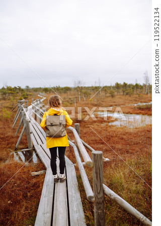 Woman traveler in a yellow coat walks along a wooden walking path along the wetlands. Back view. Woman traveler in a yellow coat walks along a wooden walking path along the wetlands. Back view. 119522134
