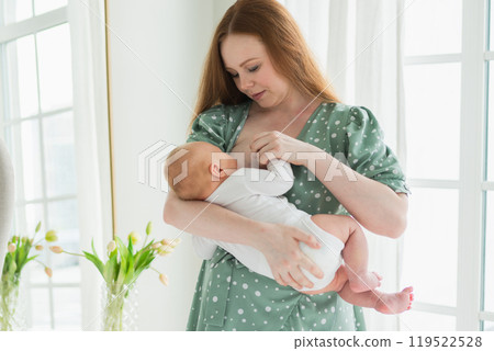 Happy family at home. Mother breastfeeding baby for health nutrition wellness. Mom nursing feeding her newborn child milk at home. Woman infant girl in white bedroom Mother hugs baby with love care 119522528