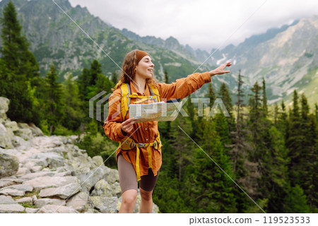 Young woman traveler with a yellow backpack, holds a map, explores hiking trails in the mountains. 119523533