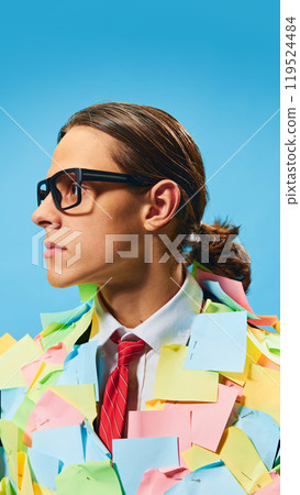 Portrait of determined young man, worker, trainee in glasses standing fully covered with colorful sticky notes against blue background. 119524484