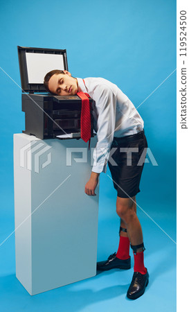 Stressed employee asleep on office copier against blue background. Young man in shorts and white shirt sleeping. Stressed employee asleep on office copier against blue background. Young man in shorts and white shirt sleeping. 119524500