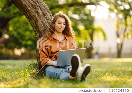 Young curly woman in the park on a green lawn with a laptop in her hands. Online education. 119524550