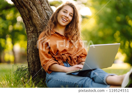 Young curly woman in the park on a green lawn with a laptop in her hands. Online education. 119524551
