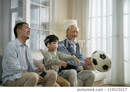 three generation asian males sitting on family couch watching live football action on tv 119525017