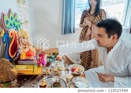 Indian couple dressed in traditional wear engages in worship at home altar, adorned with decors and offerings for god Ganesh during festive celebration. Indian culture, hindu ritual and customs. 119525499