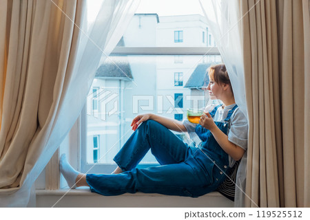 Free Time To Relax. Young woman looks out the window overlooking the city, sits on the windowsill at cozy home holds cup of hot tea drink. Happy calm female taking break for mental health wellbeing Free Time To Relax. Young woman looks out the window overlooking the city, sits on the windowsill at cozy home holds cup of hot tea drink. Happy calm female taking break for mental health wellbeing 119525512