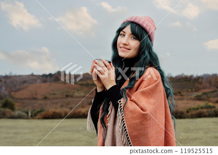 Portrait of smiling Woman in hat wrapped in Shawl Poncho drinking hot drink from thermos cup enjoying autumn landscape view. Feeling harmony, relax, personal fulfilment. Local travel lifestyle 119525515