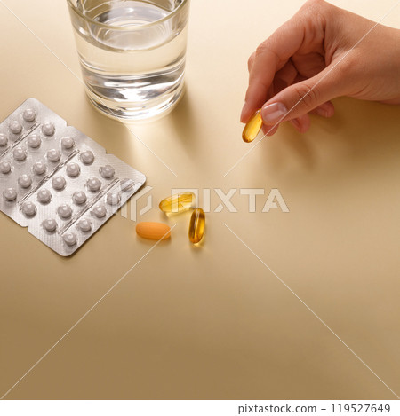 White and yellow pills, vitamins or dietary supplements lie on the table next to a glass of water, in the hand is a capsule with omega3, copy space. White and yellow pills, vitamins or dietary supplements lie on the table next to a glass of water, in the hand is a capsule with omega3, copy space. 119527649