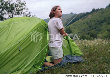 A young woman enthusiastically sets up a vibrant, green tent amidst a breathtakingly beautiful natural landscape 119527711