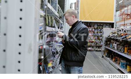Customer carefully examines power tool battery chargers while comparing products in a large hardware store. A man in a hardware store choosing a tool. 119528225