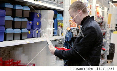 A man in a hardware store choosing a toolbox. Customer comparing plastic storage boxes in hardware store aisle, choosing best option for tools and equipment organization. Essential for home projects 119528231