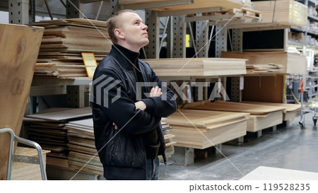 Carpenter contemplating wood options in lumber warehouse. A man in a hardware store, choosing lumber, shelves with lumber. Carpenter contemplating wood options in lumber warehouse. A man in a hardware store, choosing lumber, shelves with lumber. 119528235