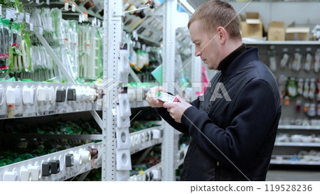 Man carefully compares electrical socket packaging on the shelf to find the best option for his needs. A man in a hardware store, choosing sockets and switches. 119528236