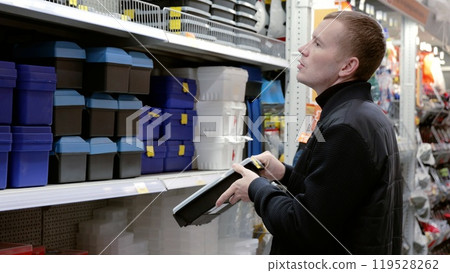 Handyman is choosing a toolbox in a hardware store, holding a black one and looking at the shelf. He is surrounded by shelves with tools and materials. A man in a hardware store choosing a tool. 119528262
