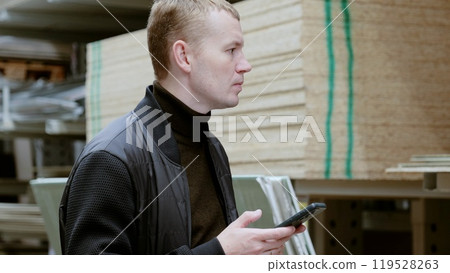 Man in a hardware store, choosing lumber, shelves with lumber. Man uses a smartphone to select plywood in a hardware store for a home construction or renovation project Man in a hardware store, choosing lumber, shelves with lumber. Man uses a smartphone to select plywood in a hardware store for a home construction or renovation project 119528263