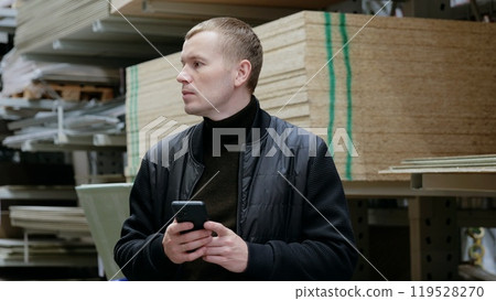 Contractor uses his smartphone to search for building material prices while in the hardware store aisle. A man in a hardware store, choosing lumber, shelves with lumber. 119528270