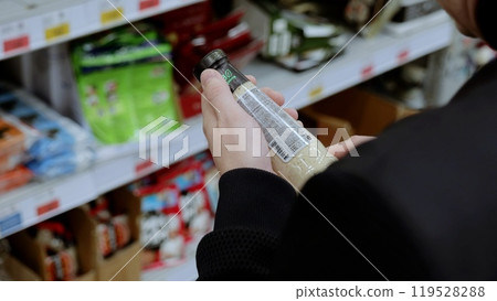 A customer in black studies the label on a bottle of sauce. Shopper carefully examines the ingredients and nutritional information on a bottle of sauce in a grocery store before buying A customer in black studies the label on a bottle of sauce. Shopper carefully examines the ingredients and nutritional information on a bottle of sauce in a grocery store before buying 119528288