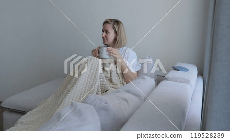 Young woman covered with a warm blanket drinks tea at home. A young woman drinks hot tea while lying on the sofa. A woman on the sofa, covered with a blanket, drinks a hot drink. Young woman covered with a warm blanket drinks tea at home. A young woman drinks hot tea while lying on the sofa. A woman on the sofa, covered with a blanket, drinks a hot drink. 119528289