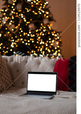 Laptop with a blank screen on a cozy sofa near a Christmas tree with festive lights in the background. Laptop with a blank screen on a cozy sofa near a Christmas tree with festive lights in the background. 119529970
