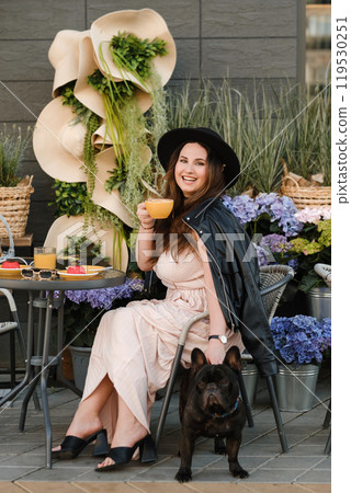 A girl is sitting in a summer cafe with her French bulldog and drinking coffee with a cake 119530251