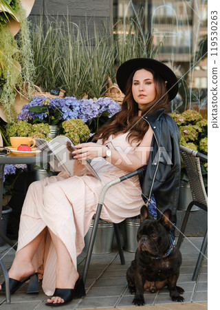 A girl is sitting in a summer cafe with her French bulldog and drinking coffee with a cake 119530263