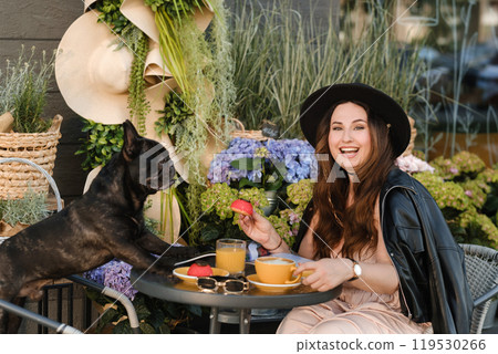 A girl is sitting in a summer cafe with her French bulldog and drinking coffee with a cake A girl is sitting in a summer cafe with her French bulldog and drinking coffee with a cake 119530266