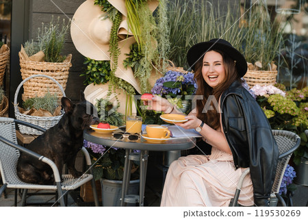 A girl is sitting in a summer cafe with her French bulldog and drinking coffee with a cake A girl is sitting in a summer cafe with her French bulldog and drinking coffee with a cake 119530269