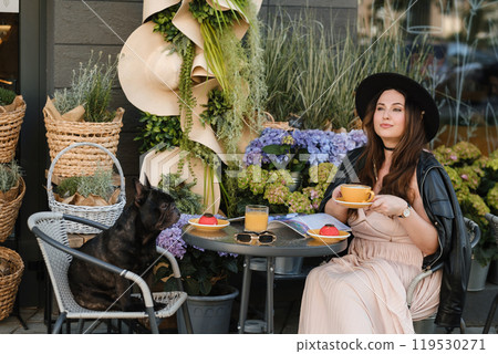 A girl is sitting in a summer cafe with her French bulldog and drinking coffee with a cake A girl is sitting in a summer cafe with her French bulldog and drinking coffee with a cake 119530271