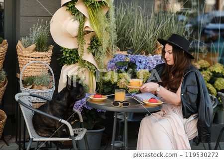 A girl is sitting in a summer cafe with her French bulldog and drinking coffee with a cake A girl is sitting in a summer cafe with her French bulldog and drinking coffee with a cake 119530272