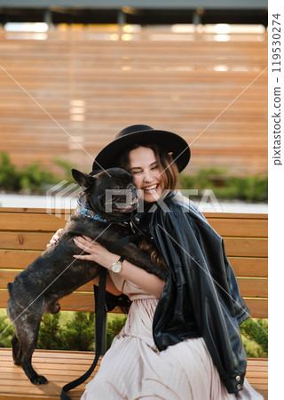 A woman in a dress and hat sits on a bench and hugs her French bulldog in the city 119530274