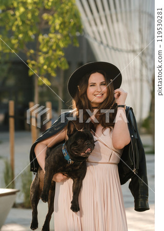 A woman in a dress and hat holds her beloved French bulldog dog in her arms against the background of the city A woman in a dress and hat holds her beloved French bulldog dog in her arms against the background of the city 119530281