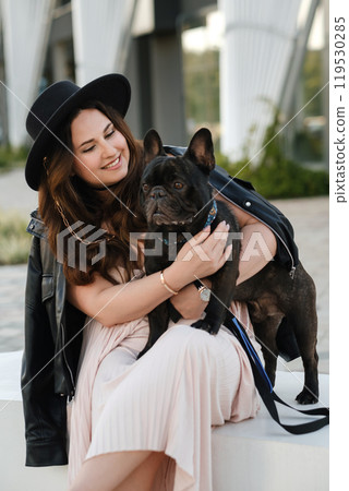 A woman in a dress and hat sits on a bench and hugs her French bulldog in the city A woman in a dress and hat sits on a bench and hugs her French bulldog in the city 119530285