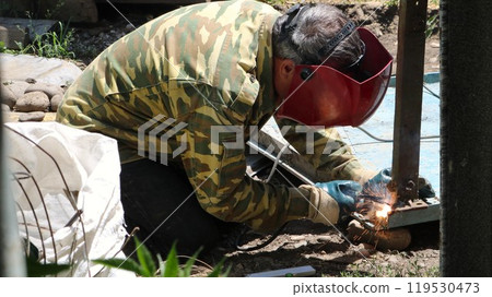 welder in red mask and green jacket works with welding tool in the process of joining two metal parts, household welding with electrodes, welding iron beams and surfaces 119530473