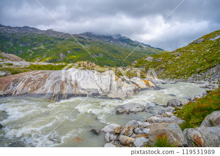 The Schlaten Glacier stream winds through the rocky landscape of Hohe Tauern National Park, surrounded by lush greenery and majestic mountains under a cloudy sky. The Schlaten Glacier stream winds through the rocky landscape of Hohe Tauern National Park, surrounded by lush greenery and majestic mountains under a cloudy sky. 119531989