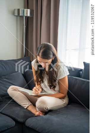 Young woman siting on the sofa and writing her personal diary, writing down thoughts, dreams and plans as part of psychotherapy after working with neurocoaching Young woman siting on the sofa and writing her personal diary, writing down thoughts, dreams and plans as part of psychotherapy after working with neurocoaching 119532067