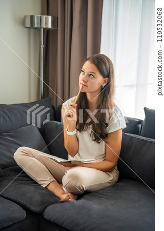 Young woman siting on the sofa and writing her personal diary, writing down thoughts, dreams and plans as part of psychotherapy after working with neurocoaching Young woman siting on the sofa and writing her personal diary, writing down thoughts, dreams and plans as part of psychotherapy after working with neurocoaching 119532068