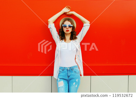 Young woman model posing wearing a casual, summer straw round hat on city street over red background Young woman model posing wearing a casual, summer straw round hat on city street over red background 119532672