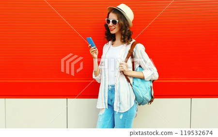 happy smiling young woman with smartphone in summer hat with backpack on red background 119532674