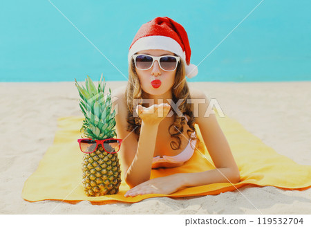 Christmas portrait woman in red santa hat with pineapple lying on beach blowing kiss on sea Christmas portrait woman in red santa hat with pineapple lying on beach blowing kiss on sea 119532704