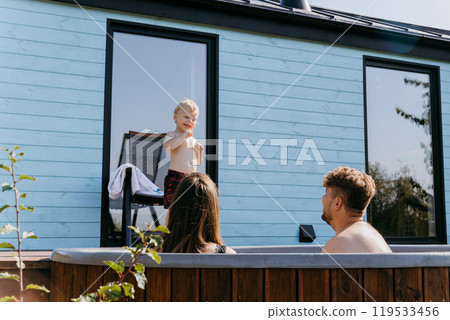 Family enjoying quality time in a backyard hot tub, with a playful child exploring the sunny day 119533456