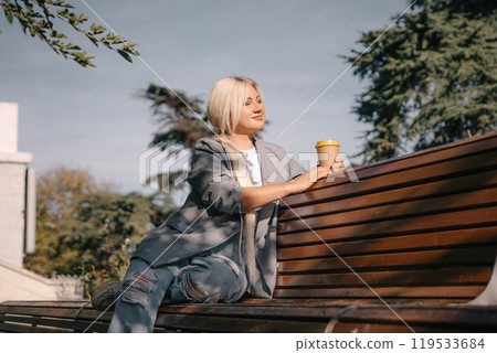 A blonde woman sits on a bench with a cup of coffee in her hand. She is wearing a gray jacket and jeans. The bench is wooden and has a curved back. The scene is peaceful and relaxing. A blonde woman sits on a bench with a cup of coffee in her hand. She is wearing a gray jacket and jeans. The bench is wooden and has a curved back. The scene is peaceful and relaxing. 119533684