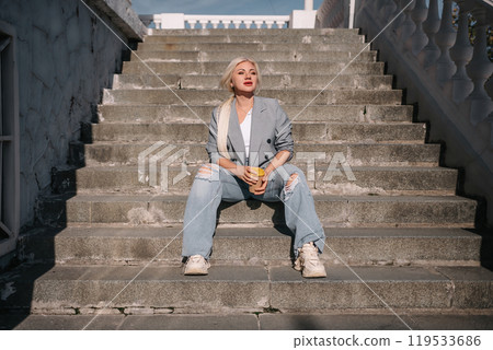 A woman in a suit and jeans sits on a set of stairs. She is holding a cup and she is smiling. A woman in a suit and jeans sits on a set of stairs. She is holding a cup and she is smiling. 119533686