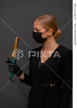 A woman in a black dress holding a hair curler. She is wearing a mask. Concept of caution and responsibility in the face of a pandemic. 119533726